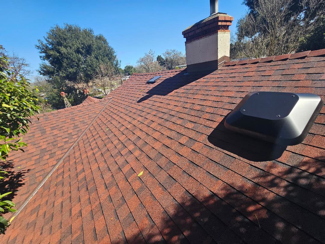 A finished roof in red blended composition shingle, photographed from
                the ridge. Courses run straight down a long hip, a brick chimney with a
                metal flue stands at the peak, a skylight sits in the slope beyond, and a
                low black roof vent is set into the field on the right. Oaks and clear
                blue sky behind.
